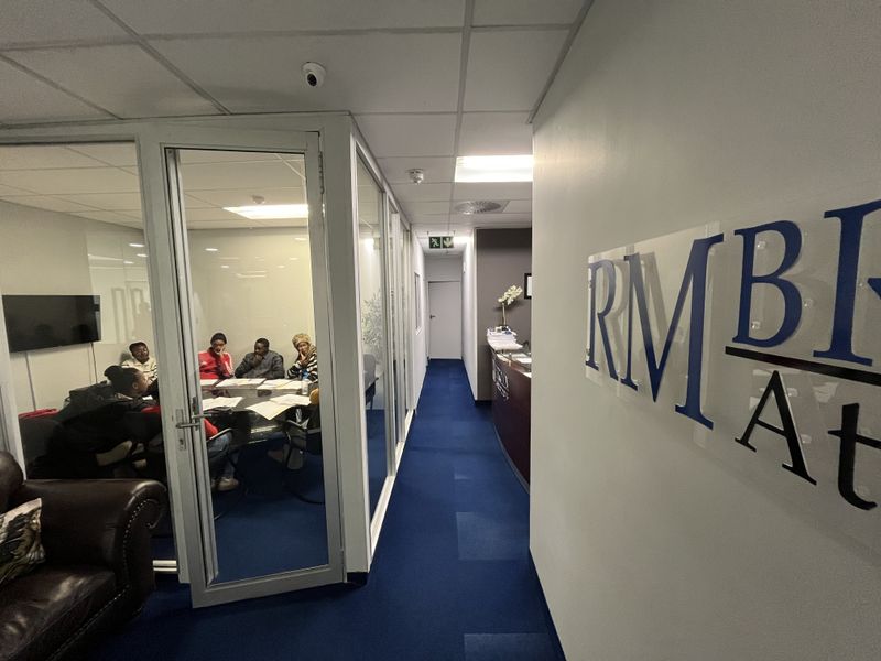 A professional office hallway featuring glass-partitioned meeting rooms, suspended ceilings with recessed lighting, and blue carpeting throughout the workspace.