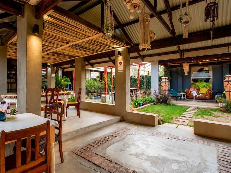 Spacious semi-outdoor restaurant patio featuring exposed timber trusses, bamboo ceiling accents, and a mix of concrete and brick flooring with integrated garden areas.