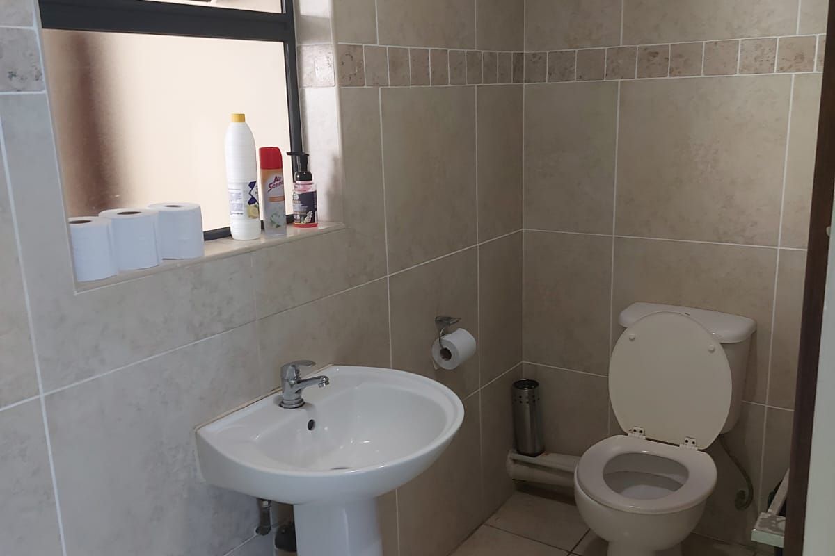 A functional bathroom featuring a pedestal sink, toilet, and neutral wall and floor tiling with a top-hung window for ventilation.