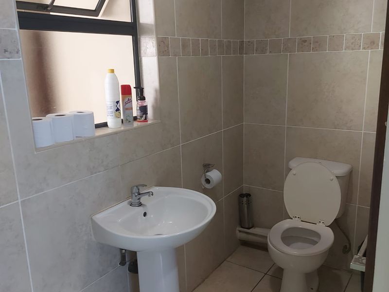 A functional bathroom featuring a pedestal sink, toilet, and neutral wall and floor tiling with a top-hung window for ventilation.