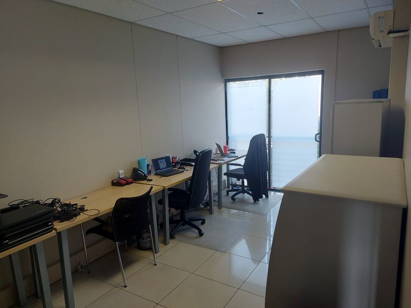 A functional office space featuring tiled flooring, a suspended ceiling with a missing tile, and a sliding glass door providing natural light.