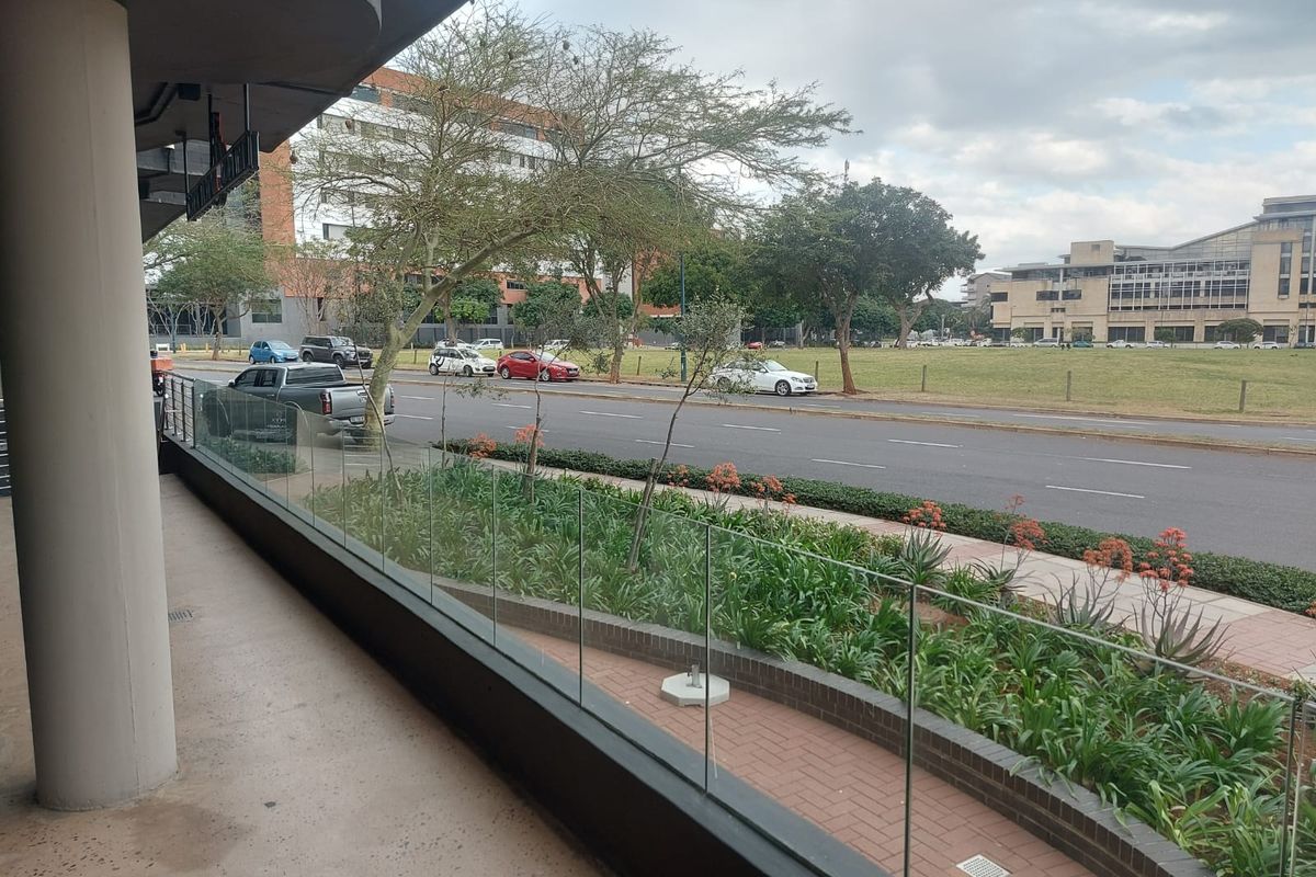 A covered outdoor balcony area featuring a modern glass balustrade, concrete structural column, and brick-paved flooring overlooking a street-side garden.
