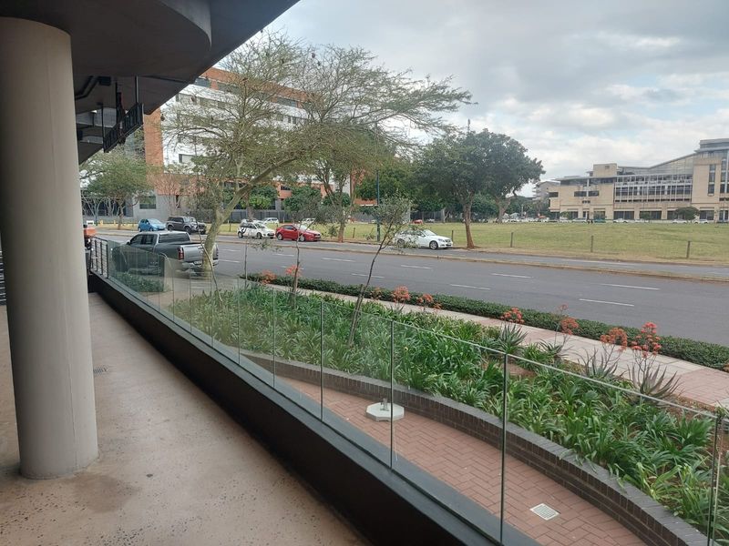 A covered outdoor balcony area featuring a modern glass balustrade, concrete structural column, and brick-paved flooring overlooking a street-side garden.