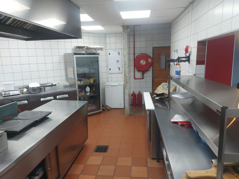 Commercial kitchen space featuring stainless steel work surfaces, tiled walls, integrated extractor hood, and essential fire safety equipment including a hose reel and extinguishers.