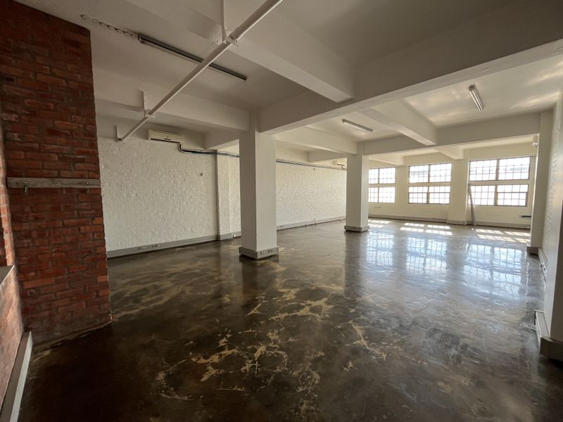 Spacious open-plan office area featuring polished concrete floors, exposed brick accents, industrial-style windows, and high ceilings with exposed structural beams.