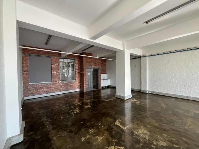 Spacious open-plan industrial office area featuring exposed brick walls, polished concrete flooring, structural beams, and a compact kitchenette unit.