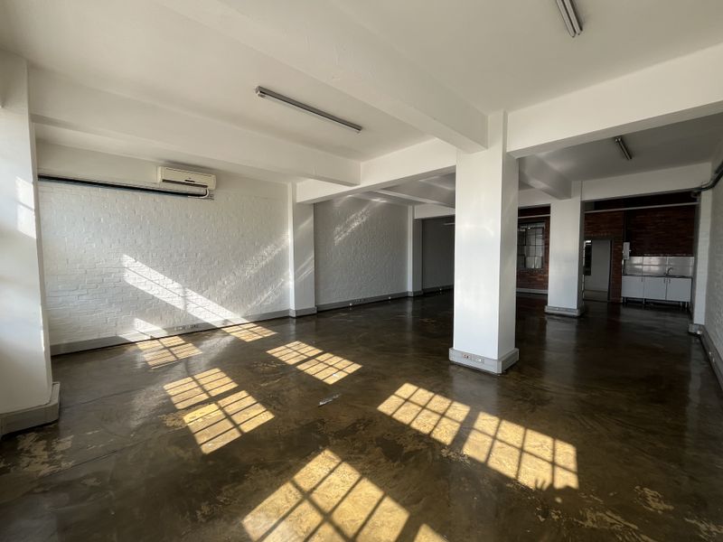 Spacious open-plan commercial area featuring polished concrete floors, exposed brick walls, structural columns, and a small kitchenette area.