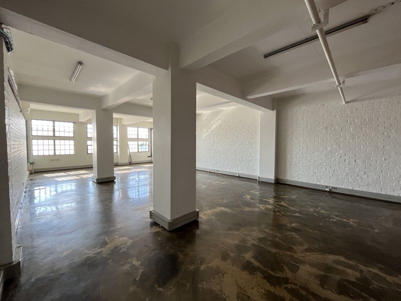Spacious open-plan industrial office area featuring polished concrete floors, exposed brick walls, and large steel-framed windows providing ample natural light.