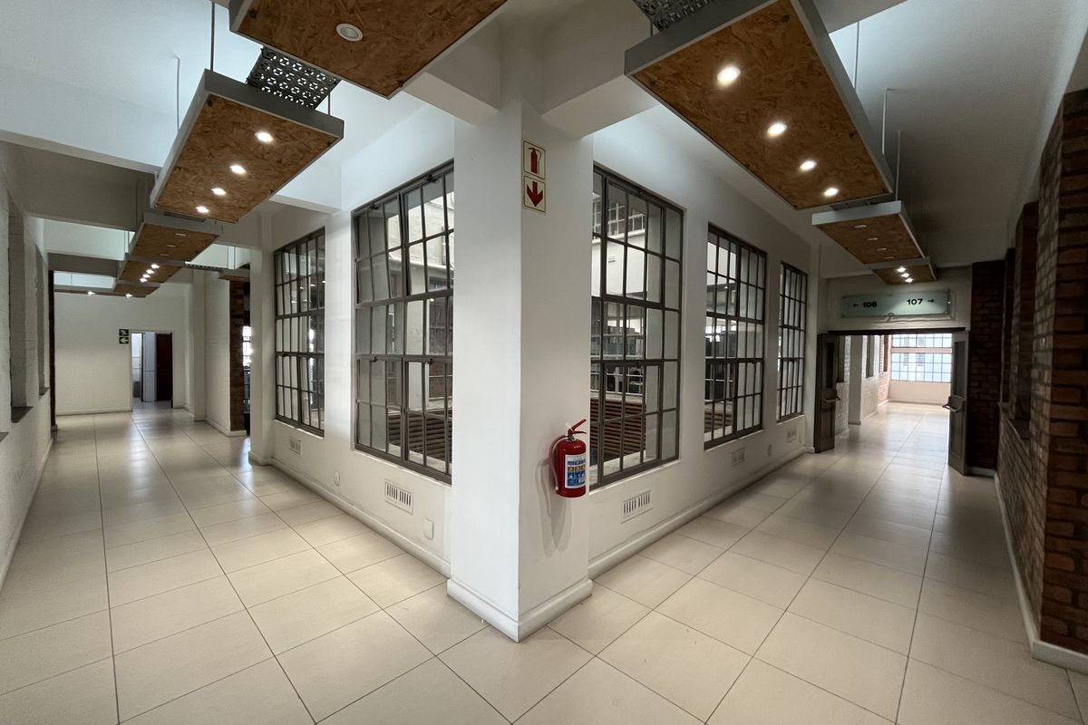 Spacious industrial-style office hallway featuring large steel-framed windows, exposed brick accents, and modern suspended lighting fixtures.