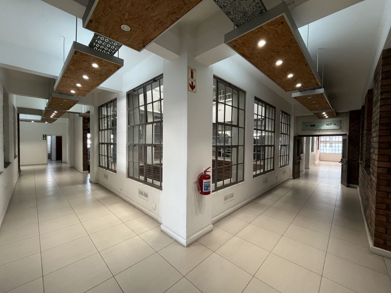 Spacious industrial-style office hallway featuring large steel-framed windows, exposed brick accents, and modern suspended lighting fixtures.