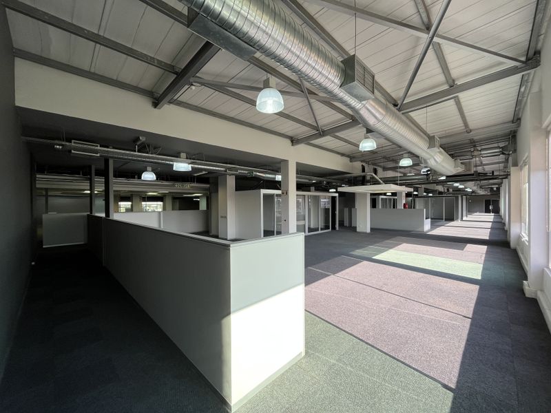Spacious open-plan office area featuring exposed industrial ceiling ducting, steel trusses, modular partition walls, and carpeted flooring throughout.