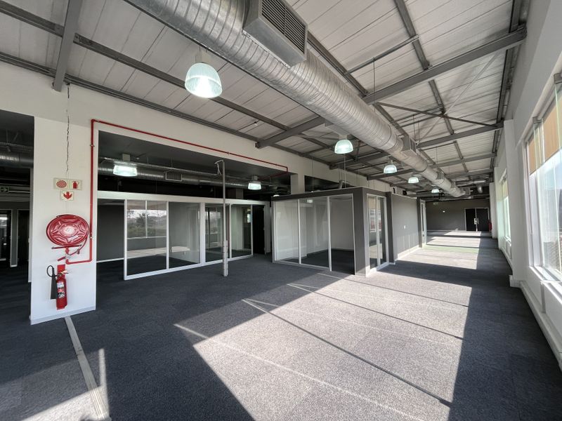 Spacious open-plan office area featuring exposed industrial ductwork, glass-partitioned meeting rooms, carpeted flooring, and abundant natural light from large windows.