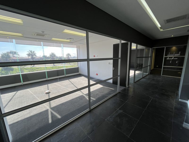 Modern vacant office space featuring glass partition walls, large windows with natural light, recessed ceiling lighting, and a combination of tiled and carpeted flooring.