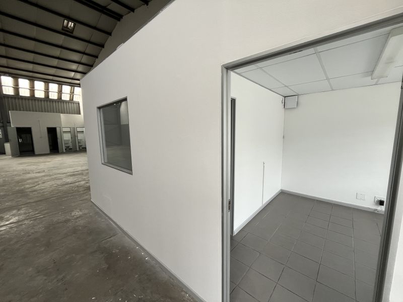 A clean, empty office space within an industrial warehouse, featuring tiled flooring, a suspended ceiling, and an internal viewing window.