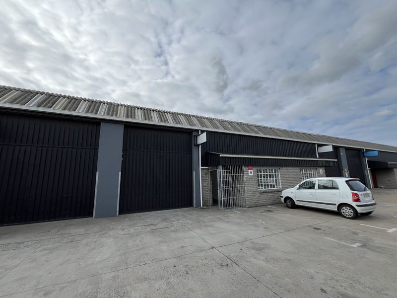 Front elevation of an industrial warehouse unit featuring dark metal cladding, roller shutter doors, and a secure entrance gate with concrete forecourt.
