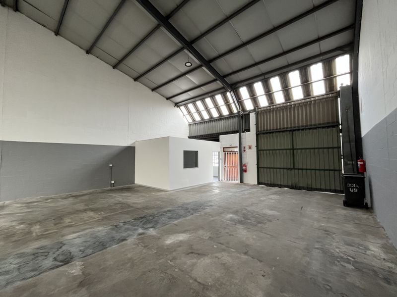 Spacious industrial warehouse unit featuring high ceilings with exposed steel trusses, concrete flooring, a large roller shutter door, and a private internal office space.