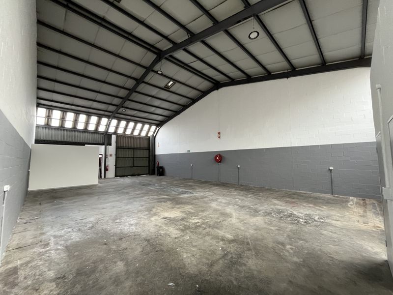 Spacious industrial warehouse unit featuring high ceilings with exposed steel trusses, concrete flooring, and a large roller shutter door for easy access.