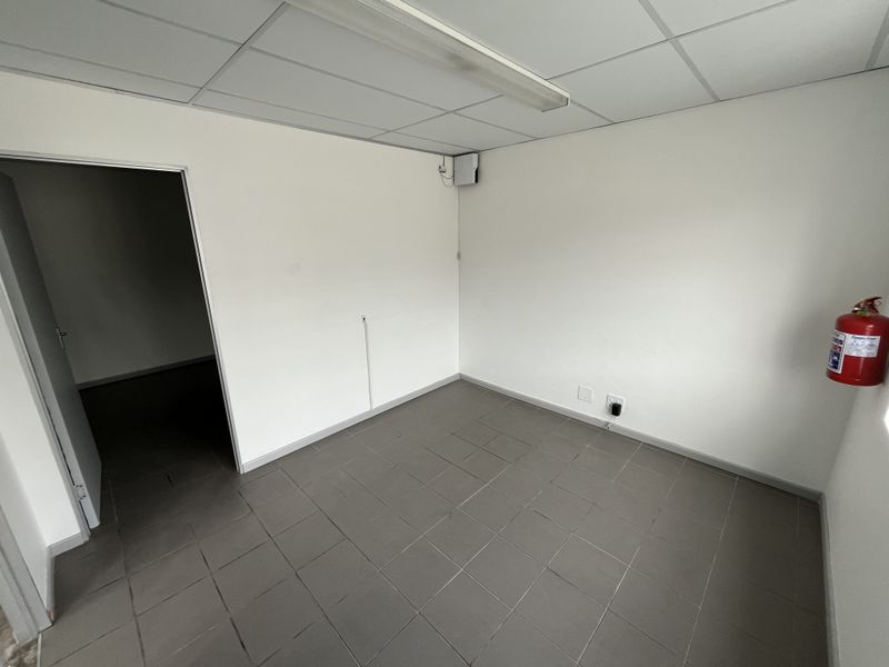 A clean, empty office space featuring neutral ceramic floor tiles, a suspended ceiling with integrated lighting, and a wall-mounted fire extinguisher.
