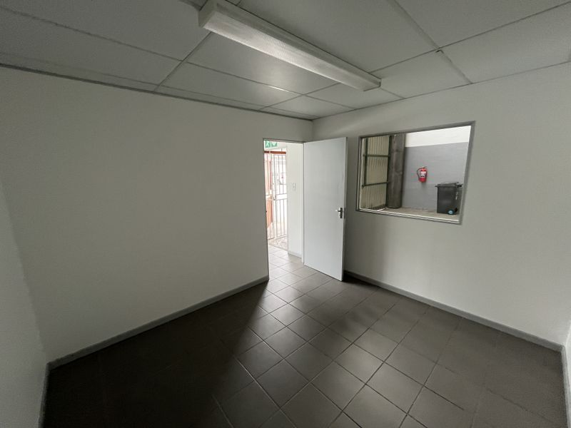 A clean, empty office space featuring tiled flooring, a suspended ceiling with integrated lighting, and an internal window looking into the industrial area.