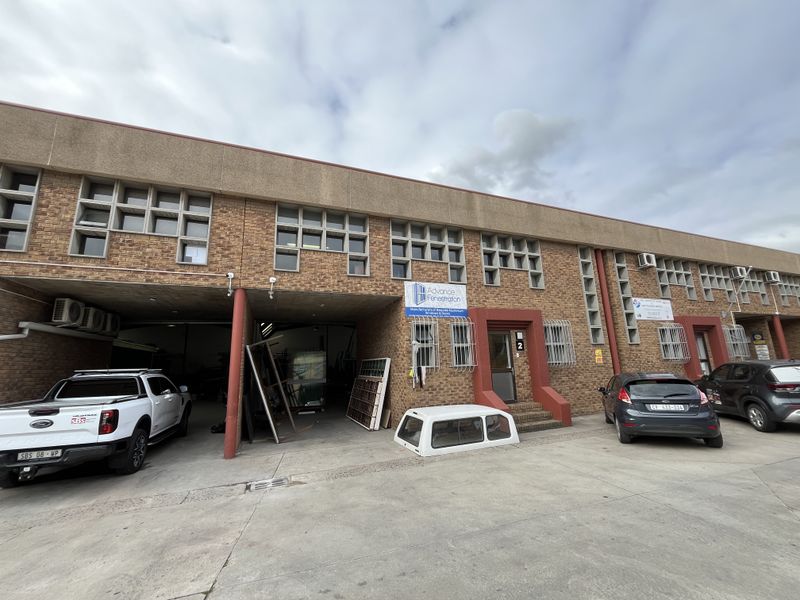 Front elevation of an industrial warehouse unit featuring brick facade, multiple access points, security bars, and dedicated concrete driveway space.