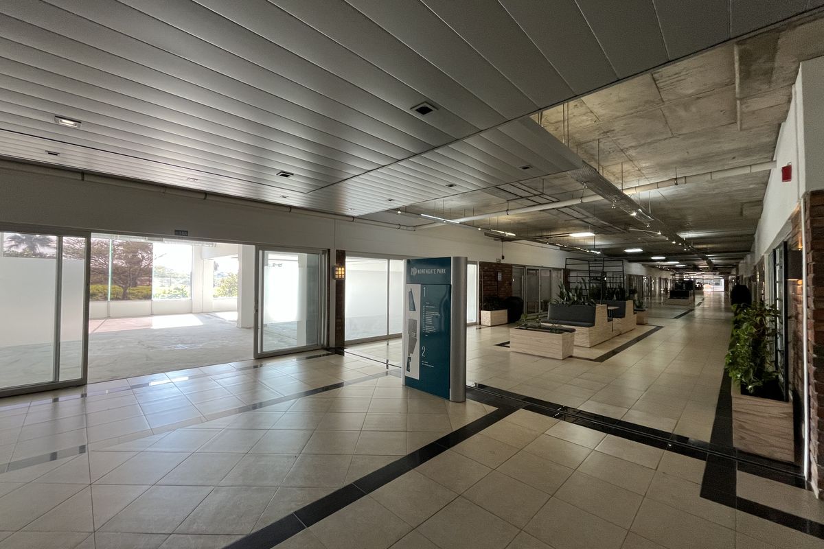 Spacious commercial hallway featuring tiled flooring, a mix of suspended metal and exposed concrete ceilings, and large sliding glass doors providing natural light.