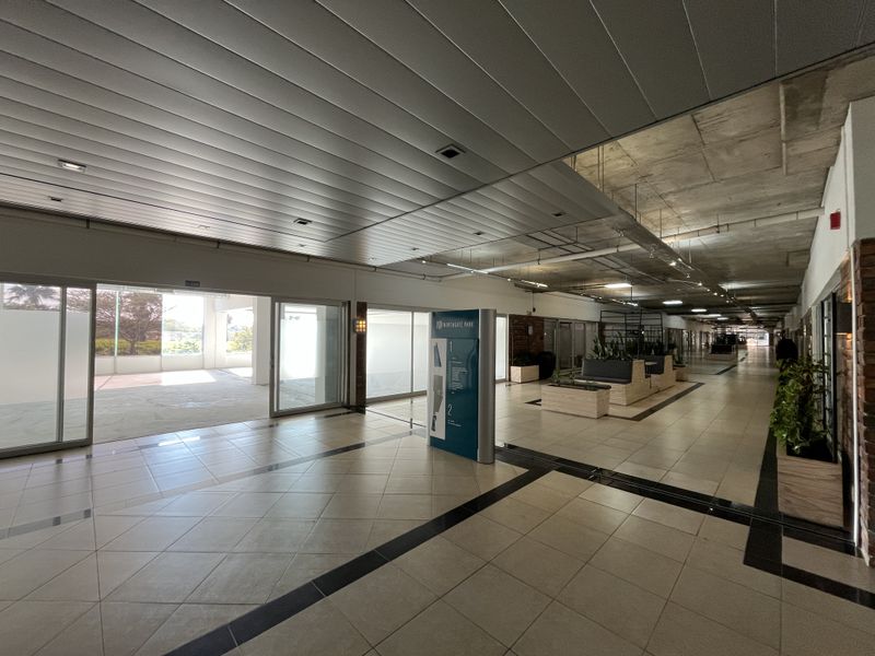 Spacious commercial hallway featuring tiled flooring, a mix of suspended metal and exposed concrete ceilings, and large sliding glass doors providing natural light.