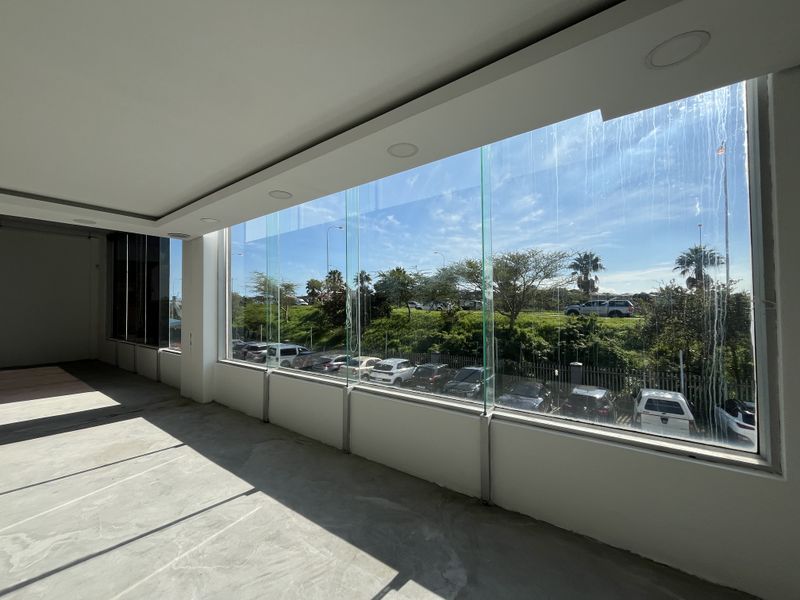 An empty, open-plan office space featuring large floor-to-ceiling windows with views of the parking area and recessed ceiling lighting.