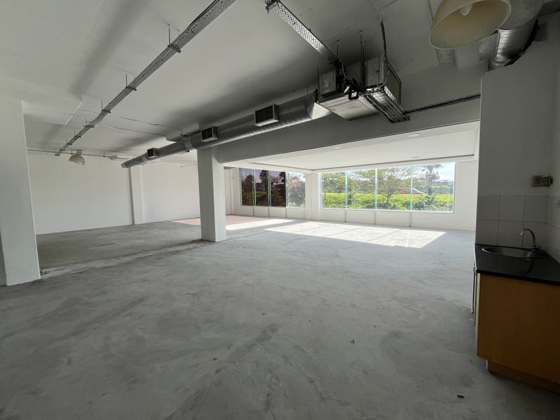 Spacious open-plan commercial area featuring exposed industrial ducting, large windows with natural light, and a basic kitchenette unit.