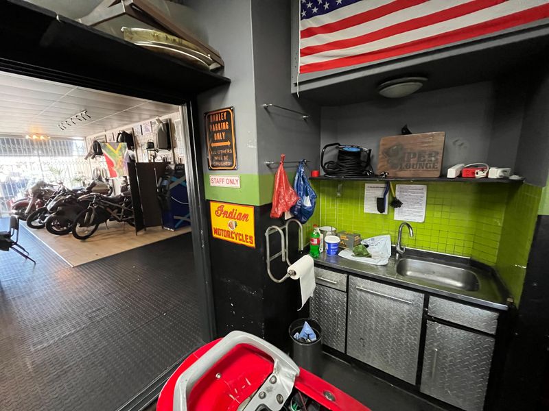 A utility scullery area featuring a stainless steel sink, diamond plate metal cabinetry, and vibrant green tiled splashback, adjacent to a workshop space.