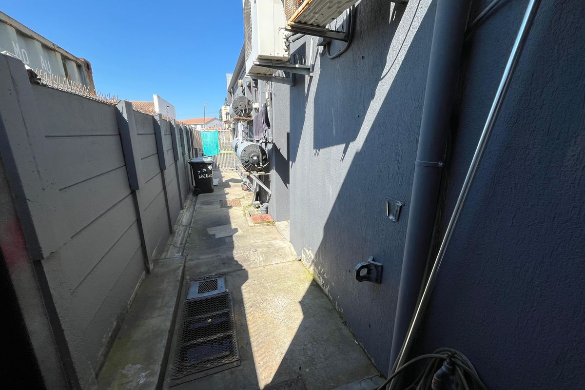 A narrow rear exterior service alley featuring concrete paving, boundary walls, and mounted air conditioning units with visible signs of wear.