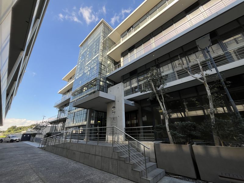 Modern multi-story office building featuring a glass curtain wall, concrete architectural elements, and stainless steel railings at the main entrance.