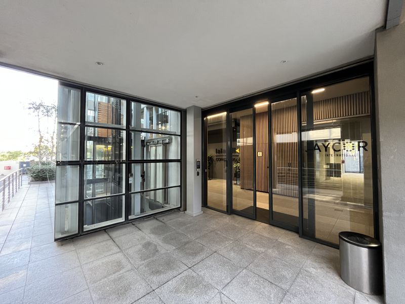 Modern office building entrance featuring a glass-enclosed elevator shaft, large sliding glass doors, and clean tiled flooring.
