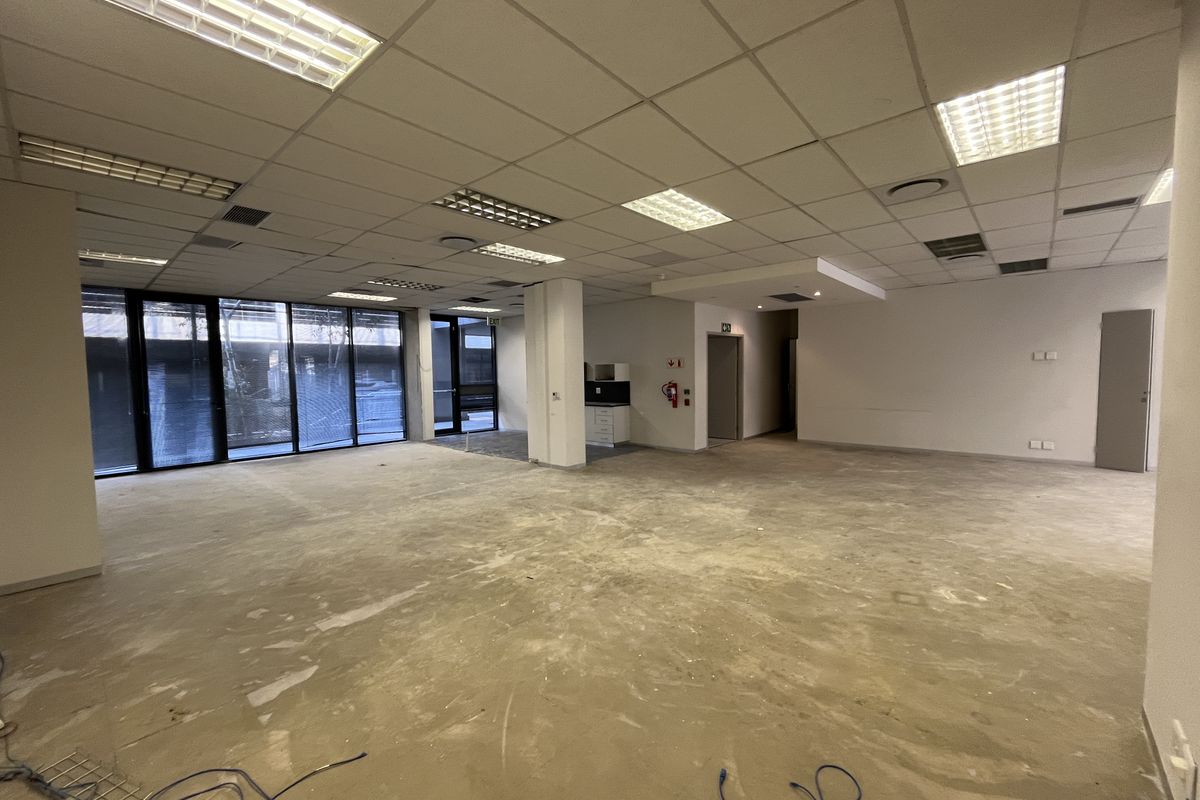 Spacious open-plan office area featuring large floor-to-ceiling windows, suspended ceilings with recessed lighting, and a small kitchenette, currently awaiting final flooring installation.