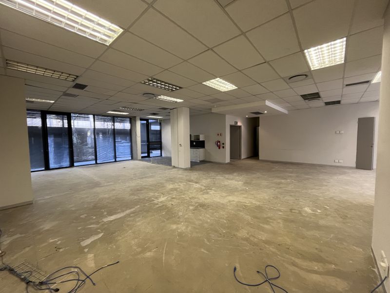 Spacious open-plan office area featuring large floor-to-ceiling windows, suspended ceilings with recessed lighting, and a small kitchenette, currently awaiting final flooring installation.