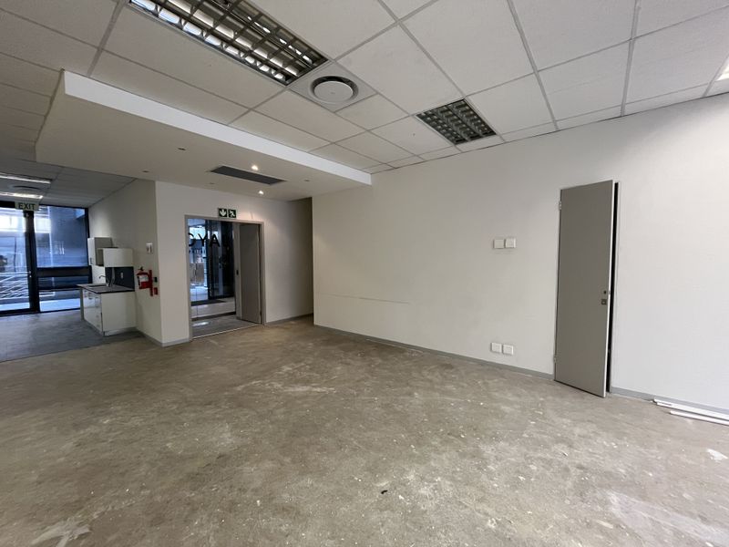 An empty office space featuring a suspended ceiling, recessed lighting, and a small kitchenette area, currently requiring flooring and wall finishing.