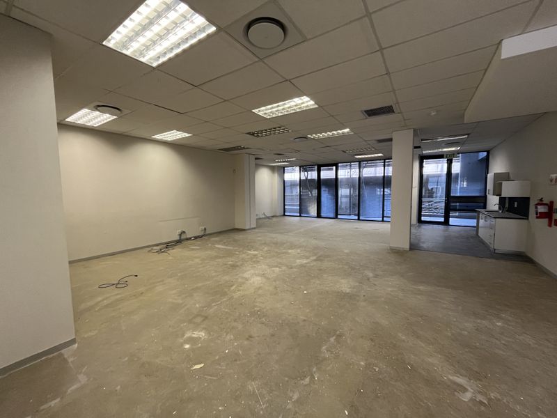 An open-plan office space featuring a suspended ceiling, recessed lighting, floor-to-ceiling windows, and a small kitchenette area, currently awaiting floor finishing.