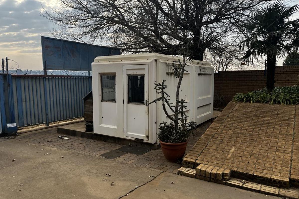 A small prefabricated security hut situated on a paved industrial yard with brick retaining walls and perimeter fencing.