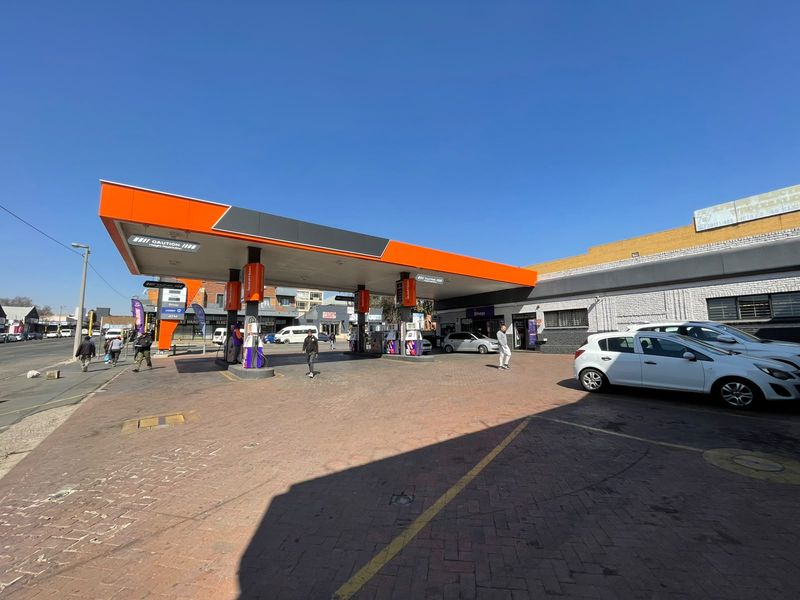 A commercial fuel station forecourt featuring a modern canopy, multiple fuel pumps, and a paved driveway area with adjacent retail shop access.