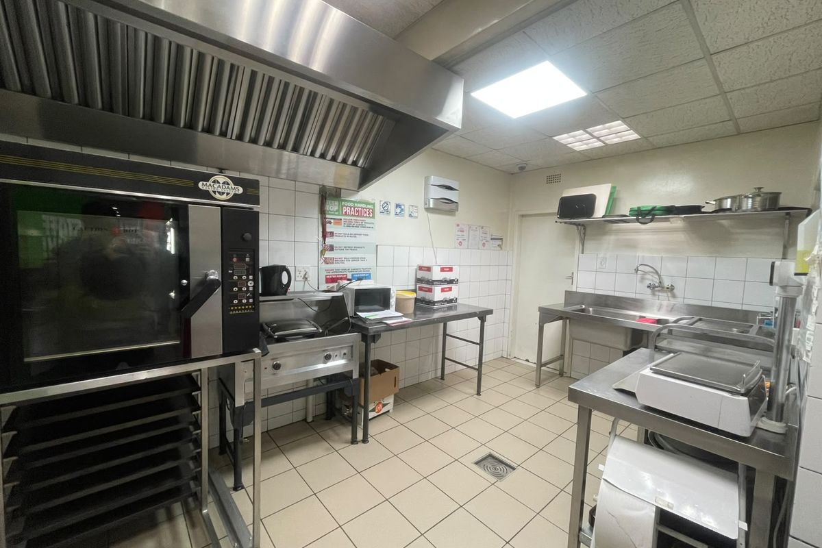 A functional commercial kitchen featuring stainless steel prep surfaces, a large extractor canopy, commercial oven, and tiled finishes with a floor drain.