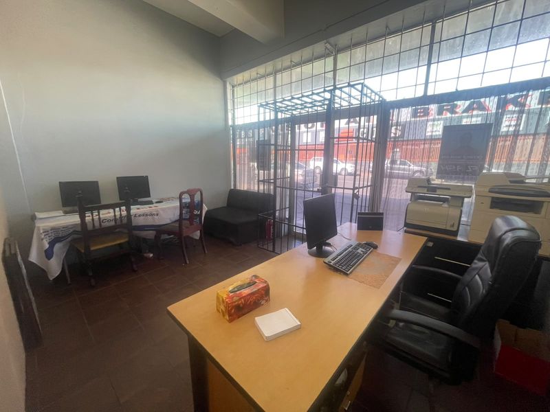 A functional retail office space featuring tiled flooring, large storefront windows with security gates, and ample natural light.