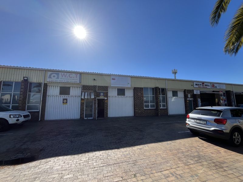 Front elevation of an industrial warehouse complex featuring multiple roller shutter doors, brickwork facade, and paved parking area.