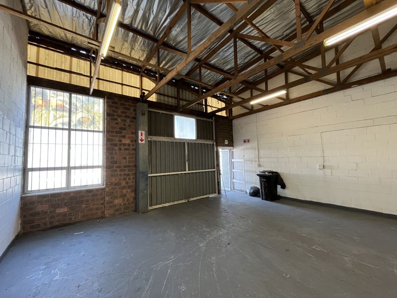 Spacious industrial utility area featuring exposed timber roof trusses, concrete flooring, large windows with security bars, and a functional roller shutter door.