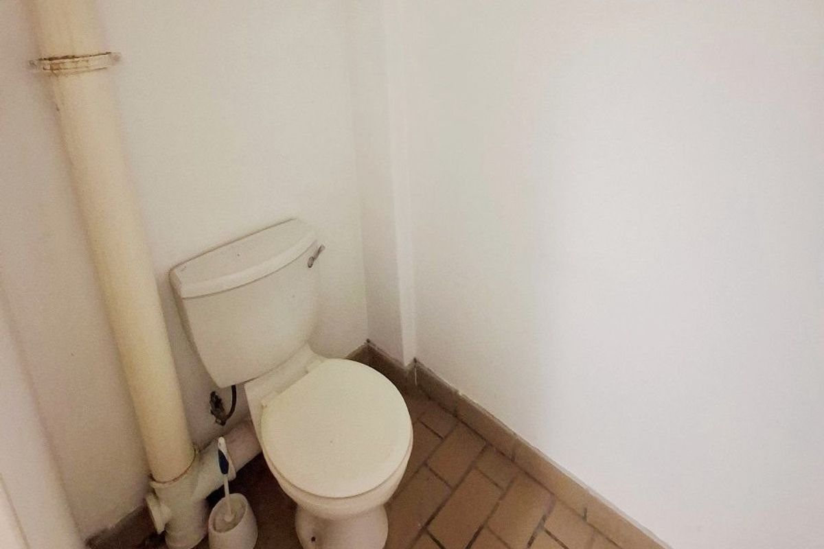 A small, basic bathroom featuring a standard toilet, exposed plumbing, and tiled flooring, currently in need of cosmetic updates and maintenance.