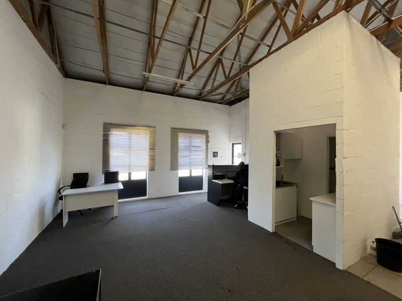 Open-plan industrial office space featuring exposed timber roof trusses, painted block walls, carpeted flooring, and a small integrated kitchenette area.