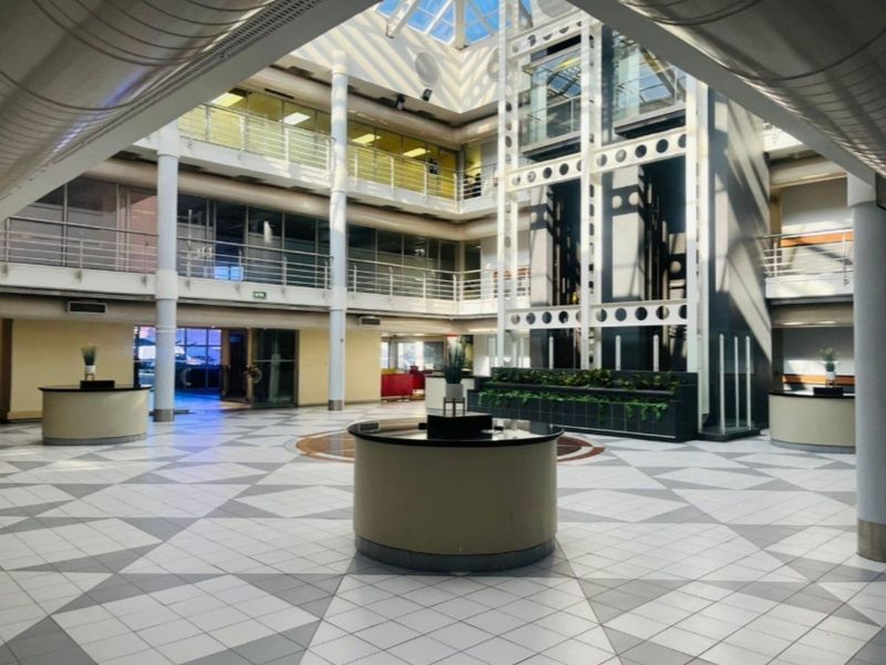 Spacious commercial atrium featuring a central glass elevator, high-reaching steel structural columns, and a large skylight providing abundant natural light.
