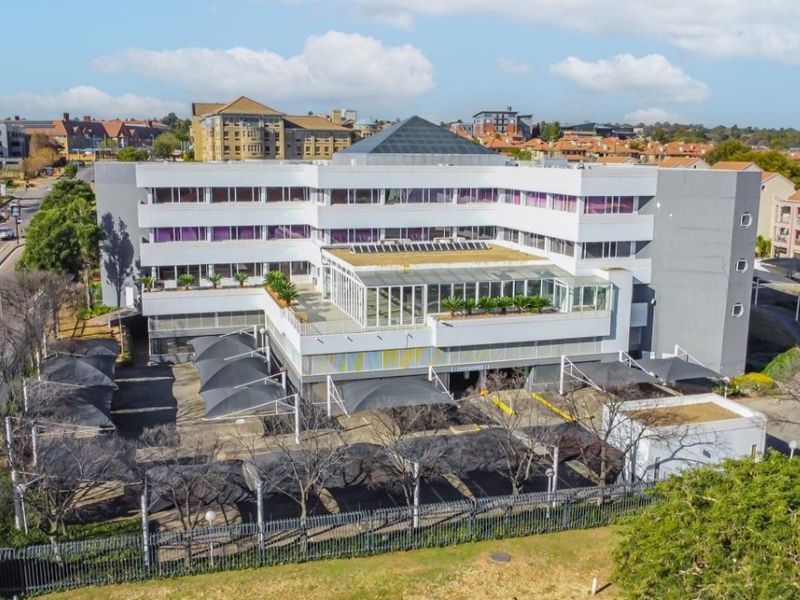 Aerial view of a multi-story commercial office building featuring a central glass atrium, extensive parking with shade sails, and perimeter security fencing.
