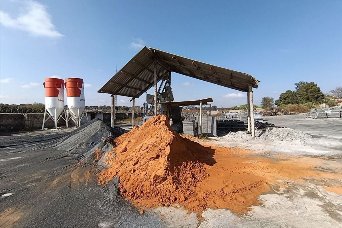 Industrial yard featuring storage silos, an open-sided metal shed structure, and large stockpiles of raw materials on a concrete surface.