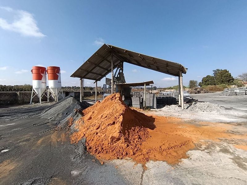 Industrial yard featuring storage silos, an open-sided metal shed structure, and large stockpiles of raw materials on a concrete surface.