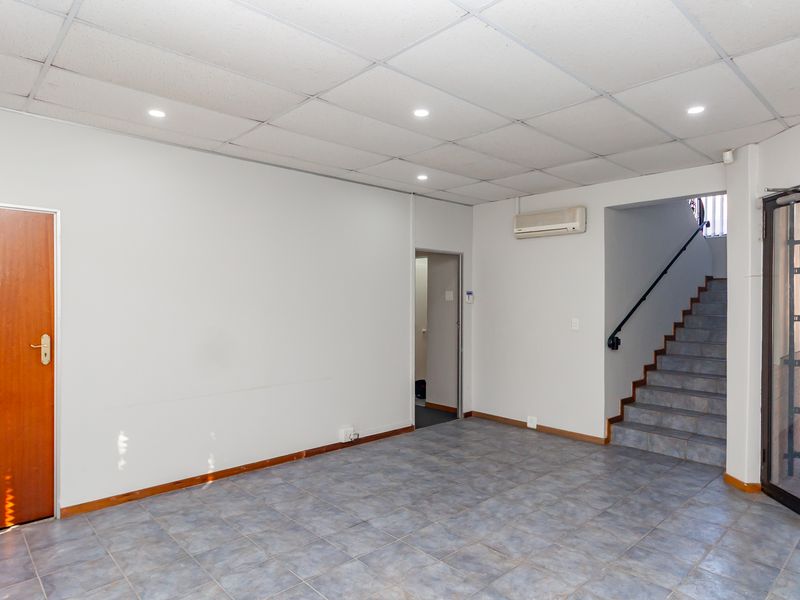 An open-plan office space featuring tiled flooring, a suspended ceiling with recessed lighting, a wall-mounted air conditioning unit, and internal staircase access.