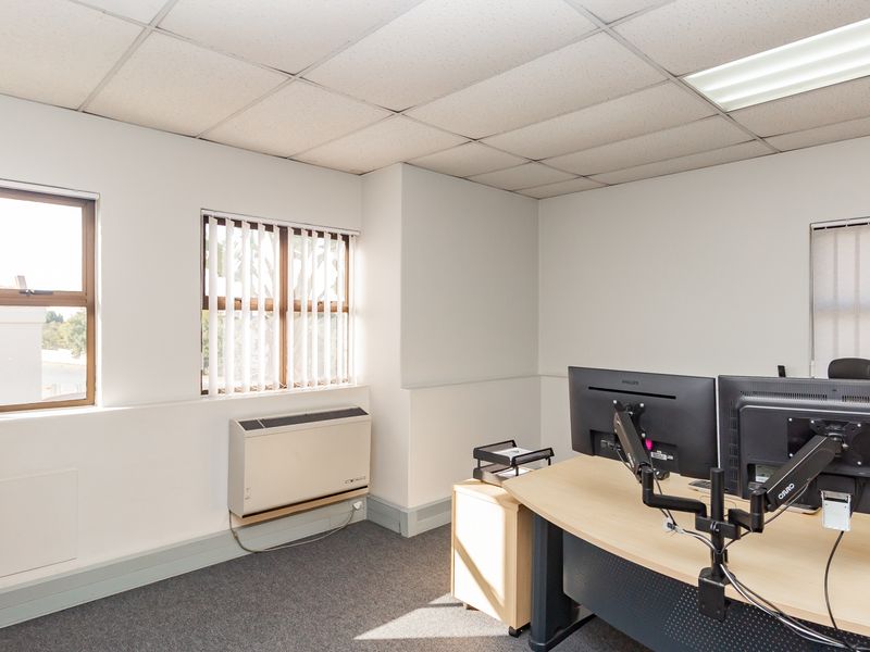 A functional office space featuring a suspended ceiling, carpeted flooring, vertical blinds, and a wall-mounted air conditioning unit.